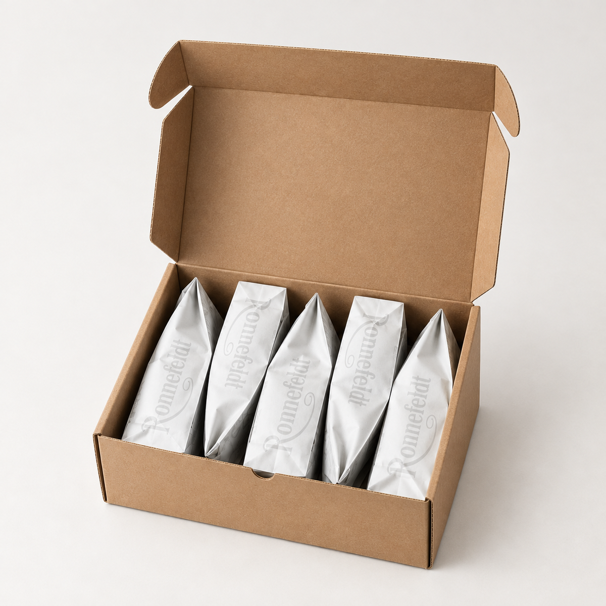An open kraft cardboard box containing five individually wrapped silver pouches, photographed on a clean light gray studio background.